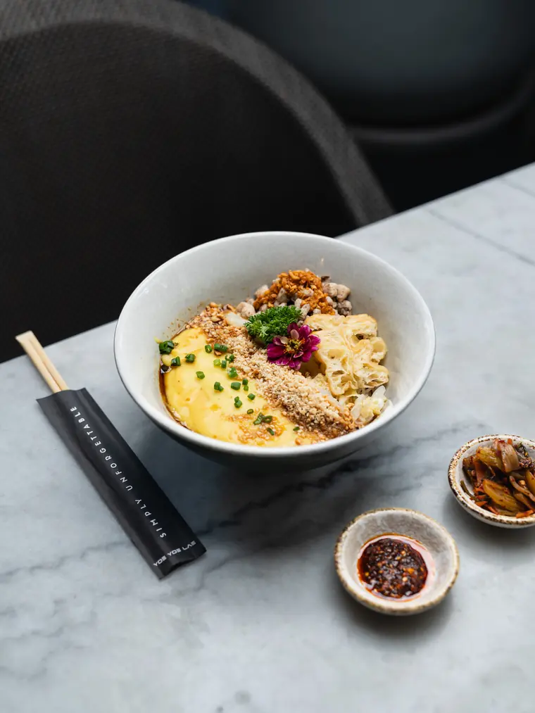 A noodle bowl on a marble table beside chilli oil and pickles.