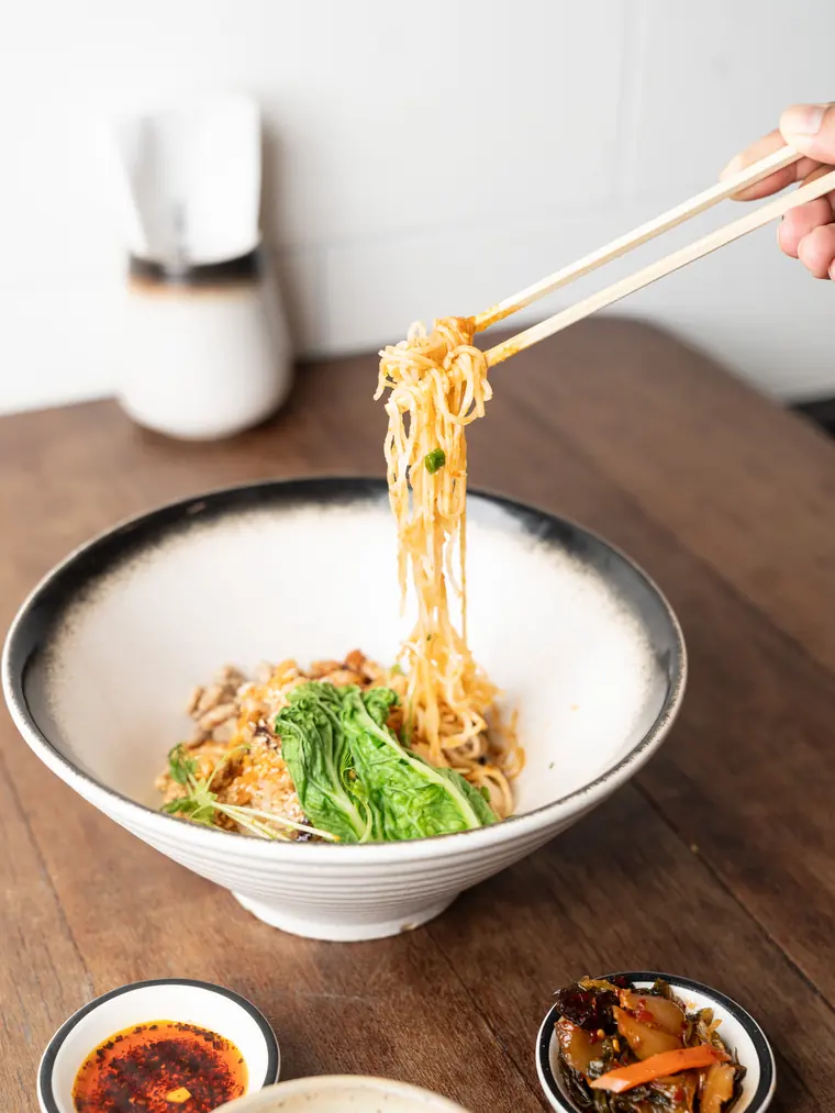 A hand lifting noodles from a broth bowl with chopsticks.