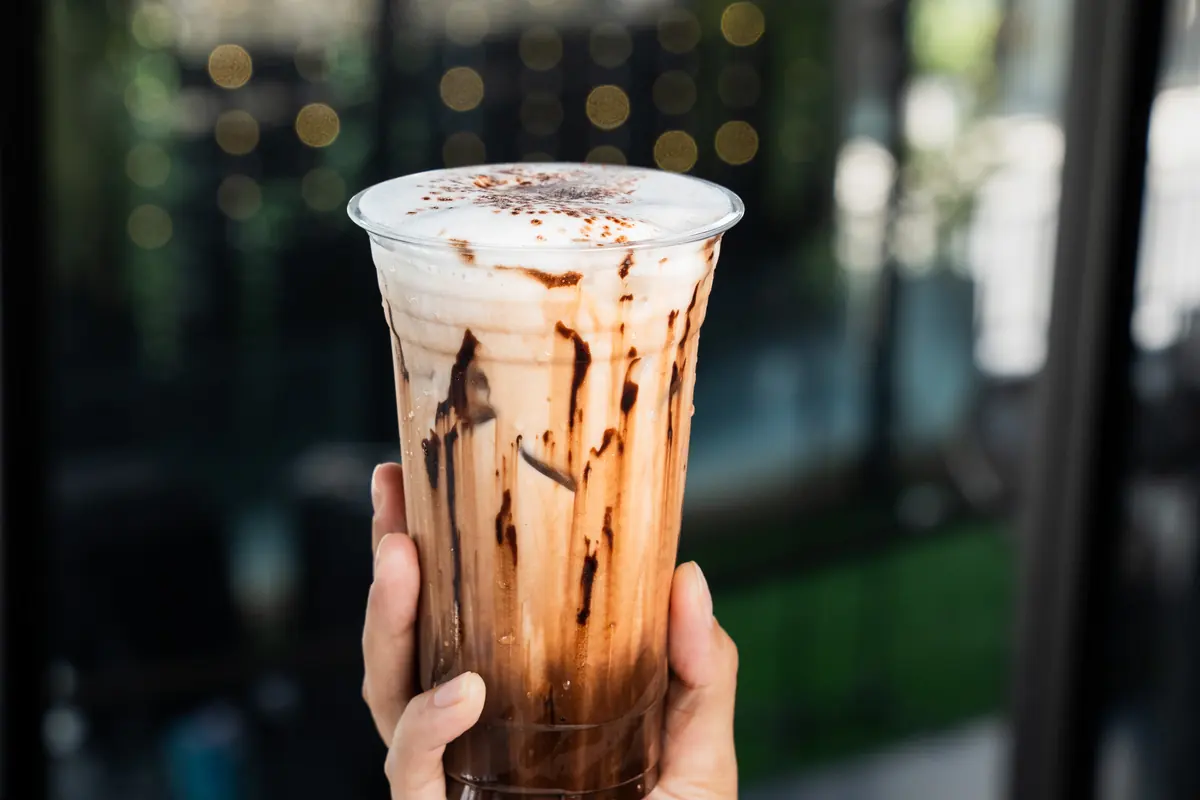 A hand holding an iced drink up to a window: a plain clear takeaway cup, chocolate drizzled down the inside under a cap of milk foam.