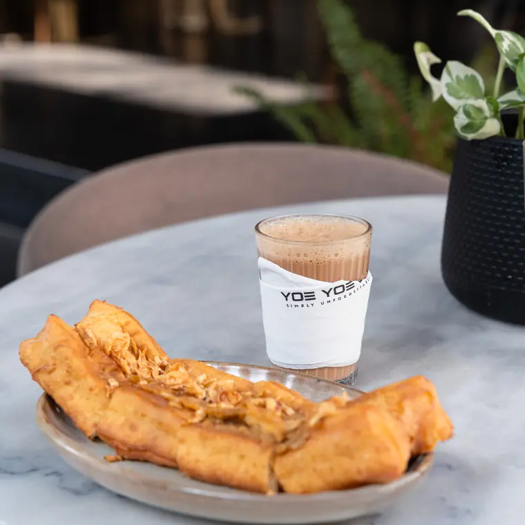 Tea in a Yoe Yoe Lae cup beside a plate of fried pastries.