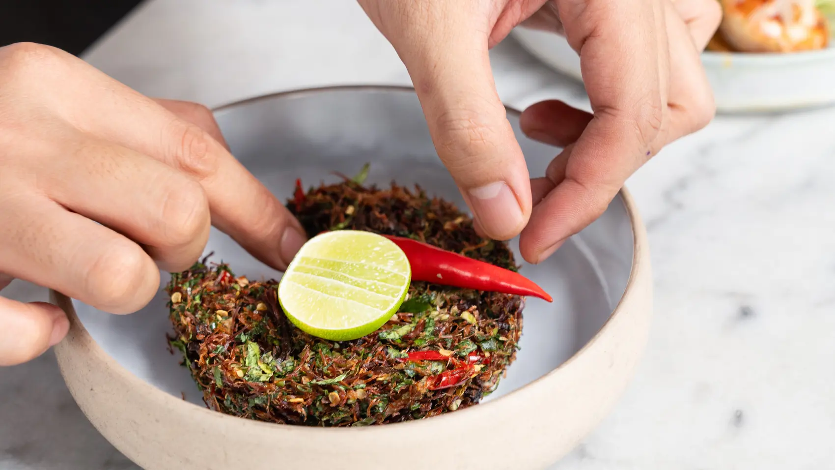Two hands placing a lime and a red chilli onto a tea-leaf salad.